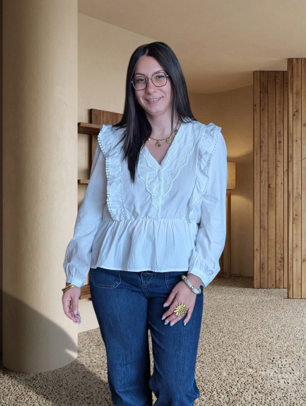 Femme portant une blouse blanche La Roberie en coton avec détails à volants et dentelle devant un mur végétal